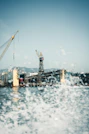 Cranes are positioned above a large industrial shipyard, with a dock and a partially visible ship. Water sprays in the foreground, adding a dynamic effect to the scene.