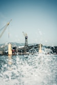 Cranes are positioned above a large industrial shipyard, with a dock and a partially visible ship. Water sprays in the foreground, adding a dynamic effect to the scene.