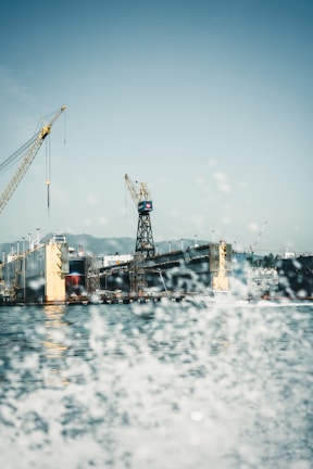Cranes are positioned above a large industrial shipyard, with a dock and a partially visible ship. Water sprays in the foreground, adding a dynamic effect to the scene.
