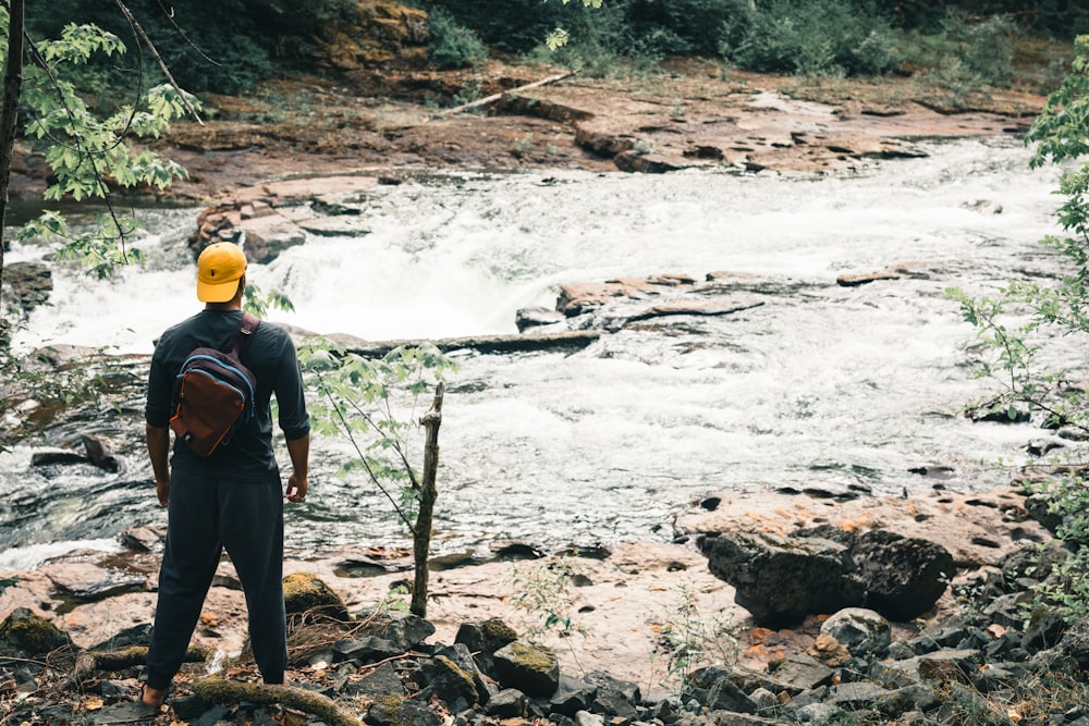 a man wearing a yellow hat standing on rocks near a river