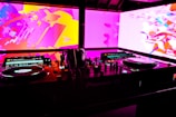 A DJ booth with technical equipment, including turntables, mixers, and headphones. The background features vibrant abstract visuals in pink, purple, and yellow hues on large screens.