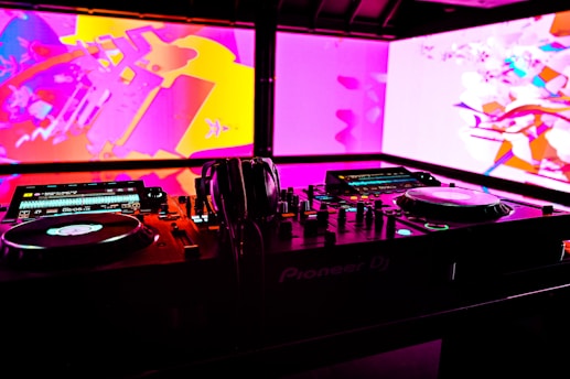 A DJ booth with technical equipment, including turntables, mixers, and headphones. The background features vibrant abstract visuals in pink, purple, and yellow hues on large screens.