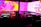 A DJ booth with technical equipment, including turntables, mixers, and headphones. The background features vibrant abstract visuals in pink, purple, and yellow hues on large screens.