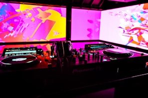 A DJ booth with technical equipment, including turntables, mixers, and headphones. The background features vibrant abstract visuals in pink, purple, and yellow hues on large screens.