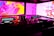 A DJ booth with technical equipment, including turntables, mixers, and headphones. The background features vibrant abstract visuals in pink, purple, and yellow hues on large screens.