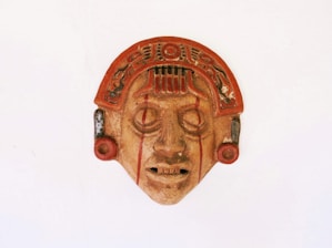 A decorative mask featuring intricate details and Aztec-style design elements. It has a symmetrical face with a prominent headdress adorned with geometric patterns, and the eyes and mouth are hollow with visible linear markings across the face.