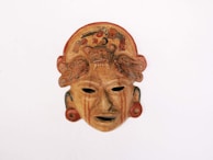 A traditional mask with intricate detailing, featuring a prominent red and brown color scheme. The mask has stylized facial features and decorative elements, including a detailed design on the forehead area.