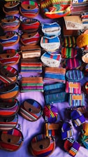 An assortment of small, colorful fabric pouches and wallets with various designs are arranged on display. Many of the items feature vibrant stripes and patterns, some include text such as 'El Salvador'. The items appear to be made of woven textiles and leather accents, showcasing craftsmanship and cultural motifs.