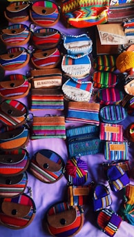 An assortment of small, colorful fabric pouches and wallets with various designs are arranged on display. Many of the items feature vibrant stripes and patterns, some include text such as 'El Salvador'. The items appear to be made of woven textiles and leather accents, showcasing craftsmanship and cultural motifs.
