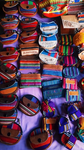 An assortment of small, colorful fabric pouches and wallets with various designs are arranged on display. Many of the items feature vibrant stripes and patterns, some include text such as 'El Salvador'. The items appear to be made of woven textiles and leather accents, showcasing craftsmanship and cultural motifs.