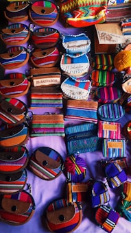 An assortment of small, colorful fabric pouches and wallets with various designs are arranged on display. Many of the items feature vibrant stripes and patterns, some include text such as 'El Salvador'. The items appear to be made of woven textiles and leather accents, showcasing craftsmanship and cultural motifs.