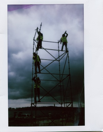 Four individuals wearing safety vests are working on a tall scaffolding against a backdrop of dark, cloudy skies. The scene appears to be an outdoor construction or setup activity near a waterfront.