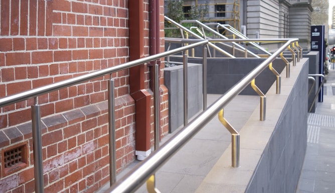 Long metal handrails are installed along a brick wall next to a sloped walkway. The wall consists of red bricks with a rectangular vent visible. The handrails are made of shiny metal and are mounted upon short posts along the edge of the walkway. Adjacent structures feature modern urban architecture.