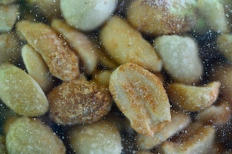 A close-up view of salted or seasoned peanuts, coated with a visible layer of salt or seasoning particles. The peanuts are clustered together, and there appears to be a slight blur or condensation effect, suggesting they are viewed through a transparent surface.