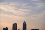 A skyline view featuring modern buildings against a pastel-colored, cloud-streaked sky. The tallest building has a distinctive triangular rooftop. A small airplane is visible in the sky.