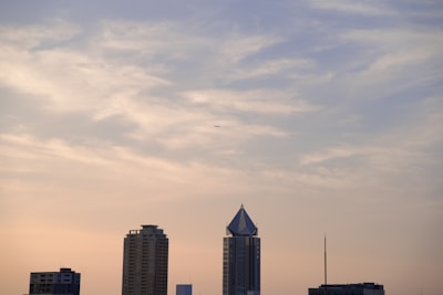 A skyline view featuring modern buildings against a pastel-colored, cloud-streaked sky. The tallest building has a distinctive triangular rooftop. A small airplane is visible in the sky.