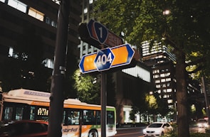 A nighttime street scene featuring a blue and orange 404 road sign prominently displayed on a pole. In the background, tall buildings with illuminated windows can be seen, along with trees lining the street. A hybrid bus and several cars are on the road, adding to the urban atmosphere.