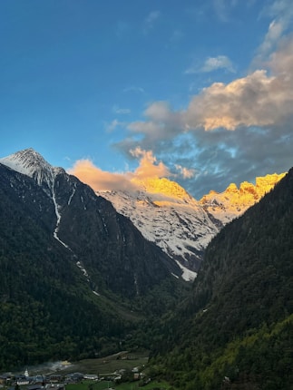 Snow-capped mountains with peaks illuminated by golden sunrise or sunset light. Dense forests cover the lower slopes, and a small village nestles in the valley below, surrounded by greenery. The sky is clear with some scattered clouds.