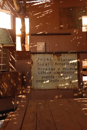 A cozy and rustic setting featuring a wooden table and walls, with dappled sunlight filtering through a wooden canopy. A vintage-looking sign displays text about local maps, photography, novels, stories, illustrated books, poetry, arts, and archaeology. A book is propped open on a shelf and a lamp with a decorative shade is visible.