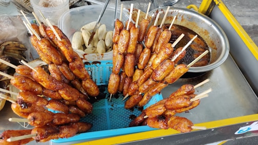 A street food vendor's cart displaying skewered, grilled bananas. Some bananas are in a stainless steel tray, marinated in a sauce, while others are neatly arranged vertically in a blue colander. The background shows utensils and containers, including peeled bananas.