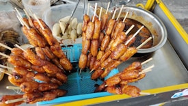 A street food vendor's cart displaying skewered, grilled bananas. Some bananas are in a stainless steel tray, marinated in a sauce, while others are neatly arranged vertically in a blue colander. The background shows utensils and containers, including peeled bananas.