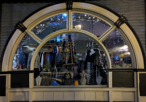 A display window of a boutique showcasing a variety of fashionable clothing and accessories. The window is elegantly designed with an arch and features decorative lights inside. Mannequins are dressed in colorful and patterned outfits, and several necklaces are displayed prominently. The reflection of the street and ambient lighting adds a cozy evening ambiance.