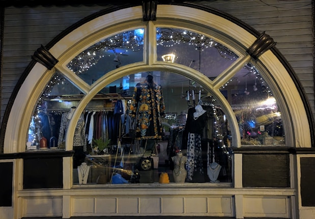 A display window of a boutique showcasing a variety of fashionable clothing and accessories. The window is elegantly designed with an arch and features decorative lights inside. Mannequins are dressed in colorful and patterned outfits, and several necklaces are displayed prominently. The reflection of the street and ambient lighting adds a cozy evening ambiance.