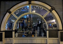 A display window of a boutique showcasing a variety of fashionable clothing and accessories. The window is elegantly designed with an arch and features decorative lights inside. Mannequins are dressed in colorful and patterned outfits, and several necklaces are displayed prominently. The reflection of the street and ambient lighting adds a cozy evening ambiance.