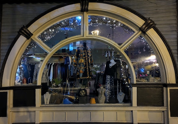 A display window of a boutique showcasing a variety of fashionable clothing and accessories. The window is elegantly designed with an arch and features decorative lights inside. Mannequins are dressed in colorful and patterned outfits, and several necklaces are displayed prominently. The reflection of the street and ambient lighting adds a cozy evening ambiance.