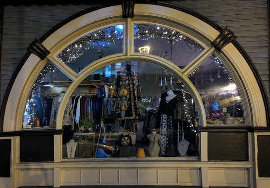 A display window of a boutique showcasing a variety of fashionable clothing and accessories. The window is elegantly designed with an arch and features decorative lights inside. Mannequins are dressed in colorful and patterned outfits, and several necklaces are displayed prominently. The reflection of the street and ambient lighting adds a cozy evening ambiance.