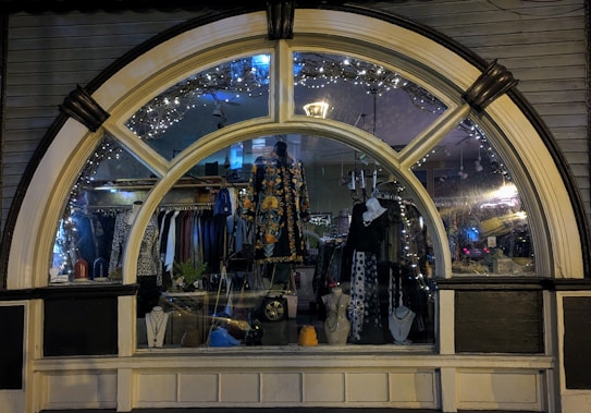 A display window of a boutique showcasing a variety of fashionable clothing and accessories. The window is elegantly designed with an arch and features decorative lights inside. Mannequins are dressed in colorful and patterned outfits, and several necklaces are displayed prominently. The reflection of the street and ambient lighting adds a cozy evening ambiance.