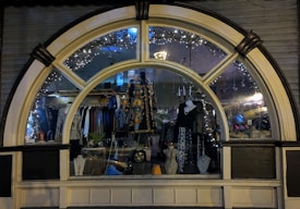 A display window of a boutique showcasing a variety of fashionable clothing and accessories. The window is elegantly designed with an arch and features decorative lights inside. Mannequins are dressed in colorful and patterned outfits, and several necklaces are displayed prominently. The reflection of the street and ambient lighting adds a cozy evening ambiance.
