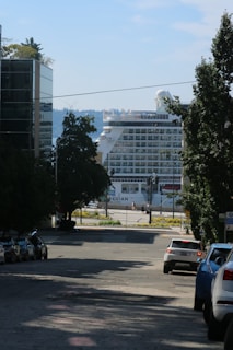 A large cruise ship is visible at the end of a city street, framed by tall trees and buildings. Several parked cars line the street. The ship's multiple decks and distinctive shape stand out against the distant backdrop of a clear blue sky and sparse treetops.
