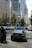 A busy city street scene with several cars and a motorcycle in traffic. In the background, tall modern buildings with reflective glass facades and a banner for the Seattle Art Museum are visible. Trees line the street, adding some greenery to the urban setting.