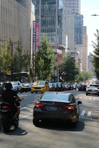 A busy city street scene with several cars and a motorcycle in traffic. In the background, tall modern buildings with reflective glass facades and a banner for the Seattle Art Museum are visible. Trees line the street, adding some greenery to the urban setting.