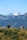 A group of five people walking on a grassy hill with scattered small trees, surrounded by majestic mountains topped with snow. The sky is clear, offering a panoramic view of the rugged terrain.