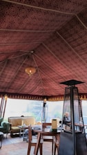 The interior of a tent with an intricately patterned maroon fabric ceiling and a warm chandelier hanging from the center. The space is furnished with a round wooden table, wooden chairs, and a cozy seating area featuring couches with cushions. A tall outdoor heater stands beside the table.