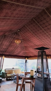 The interior of a tent with an intricately patterned maroon fabric ceiling and a warm chandelier hanging from the center. The space is furnished with a round wooden table, wooden chairs, and a cozy seating area featuring couches with cushions. A tall outdoor heater stands beside the table.