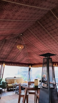 The interior of a tent with an intricately patterned maroon fabric ceiling and a warm chandelier hanging from the center. The space is furnished with a round wooden table, wooden chairs, and a cozy seating area featuring couches with cushions. A tall outdoor heater stands beside the table.