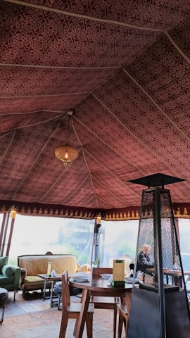 The interior of a tent with an intricately patterned maroon fabric ceiling and a warm chandelier hanging from the center. The space is furnished with a round wooden table, wooden chairs, and a cozy seating area featuring couches with cushions. A tall outdoor heater stands beside the table.