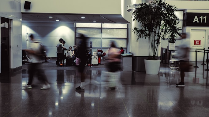 A busy airport terminal with several blurred figures walking past, suggesting a fast-paced environment. In the background, people are seated near their luggage, some engaging in conversation. The terminal features modern design elements, including large windows, overhead lighting, and a potted plant near the gate marked A11.
