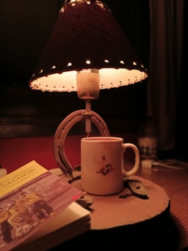 A dimly lit table with a rustic lamp featuring a dark shade and textile details. Below the lamp, there's a white mug with a bird illustration. A book titled 'Eduardo Sacheri' with sepia-toned cover art is partially visible in the foreground.