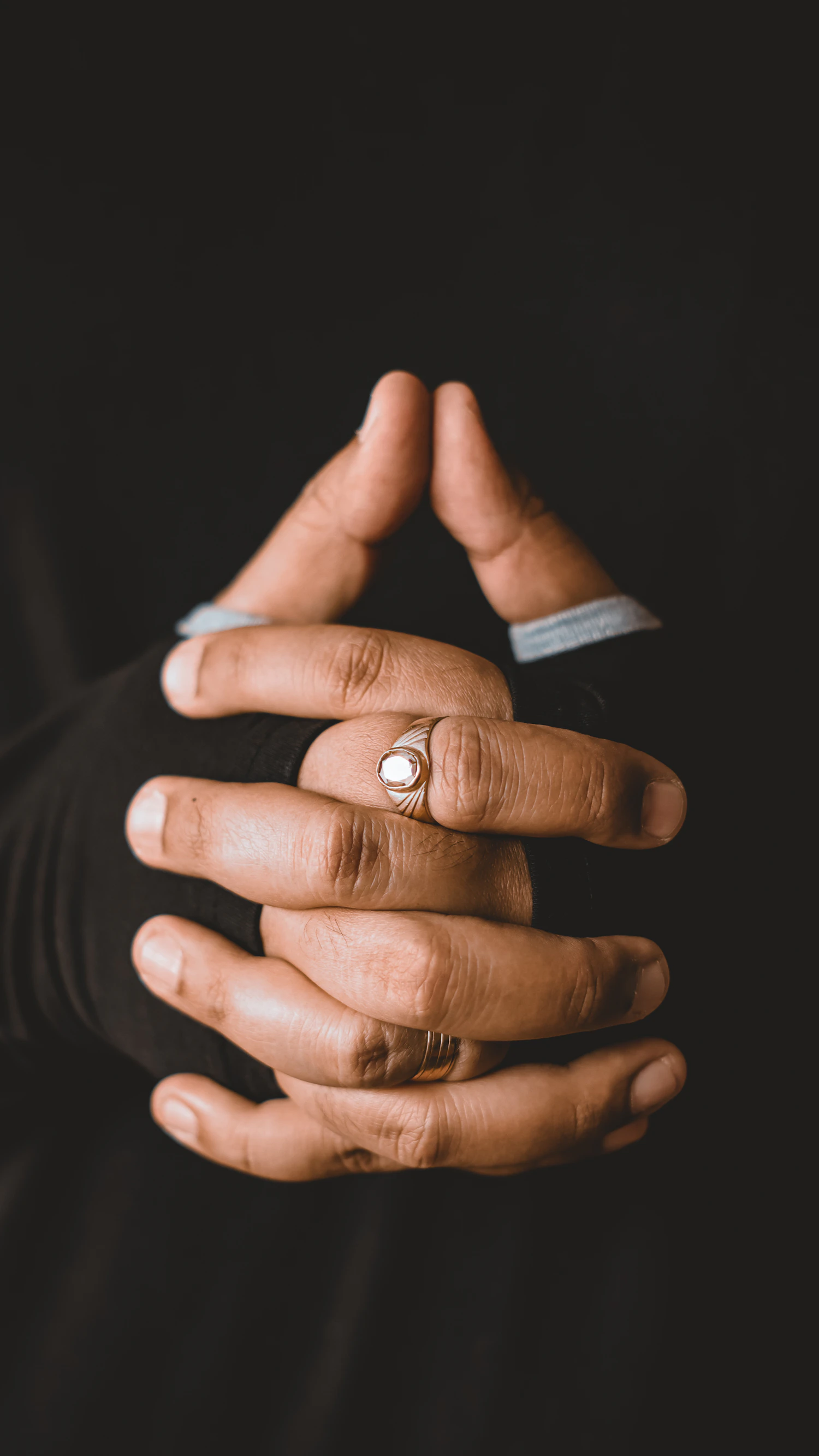 Two dark-skinned hands clasped together, each wearing a plain gold band, photographed against a dark ground