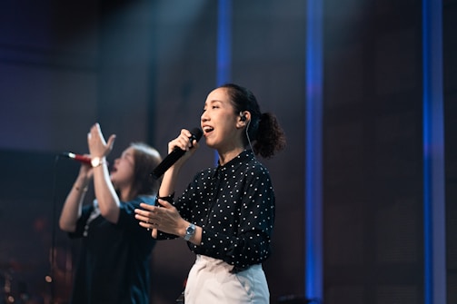 A woman is singing passionately on stage, holding a black microphone to her mouth. She is wearing a black, long-sleeve, polka-dot blouse and white pants. Her hair is tied back and she is illuminated by stage lights. In the background, another person is blurred, appearing to sing or speak with raised hands. The setting suggests a musical or performance environment with subdued lighting and a blue-toned backdrop.