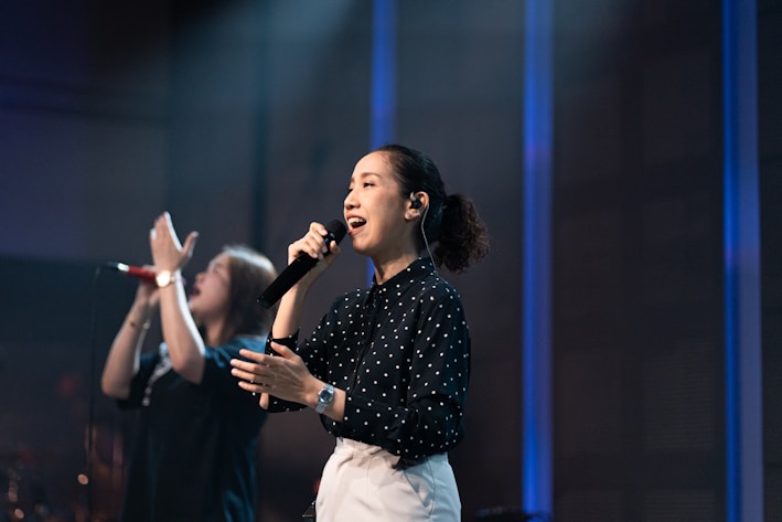 A woman is singing passionately on stage, holding a black microphone to her mouth. She is wearing a black, long-sleeve, polka-dot blouse and white pants. Her hair is tied back and she is illuminated by stage lights. In the background, another person is blurred, appearing to sing or speak with raised hands. The setting suggests a musical or performance environment with subdued lighting and a blue-toned backdrop.