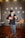 A person wearing a black hat, black jacket, and red tie is playing a ukulele while standing in front of a wall decorated with plates. A wooden table with another ukulele and some candles is also present.