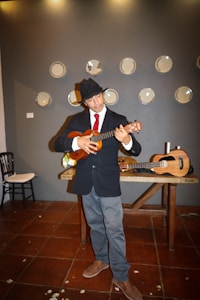 A person wearing a black hat, black jacket, and red tie is playing a ukulele while standing in front of a wall decorated with plates. A wooden table with another ukulele and some candles is also present.