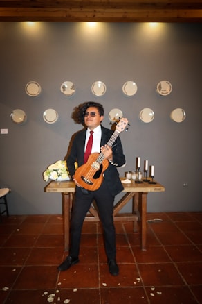 A person in formal attire holds a ukulele, standing in front of a wooden table adorned with flowers and black candles. The background features a wall decorated with plates arranged in a pattern.