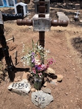 A rustic, handmade wooden cross marks a grave, surrounded by dry, brown earth. A bouquet of colorful flowers, including pink, purple, and white blossoms, is placed at the base of the cross in a glass container. Two rocks inscribed with letters 'EFSM' and 'JNS' are positioned in front. Tombstones and other crosses are visible in the background of a sunlit cemetery.
