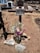A rustic, handmade wooden cross marks a grave, surrounded by dry, brown earth. A bouquet of colorful flowers, including pink, purple, and white blossoms, is placed at the base of the cross in a glass container. Two rocks inscribed with letters 'EFSM' and 'JNS' are positioned in front. Tombstones and other crosses are visible in the background of a sunlit cemetery.